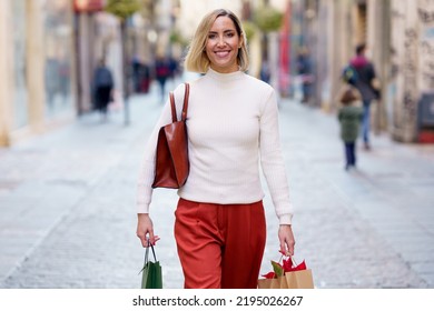 Happy female in stylish outfit carrying shopping bags with purchases and looking at camera while strolling on paved street of city - Shutterstock ID 2195026267