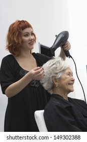 Happy Female Stylist Blow Drying Senior Woman's Hair In Salon