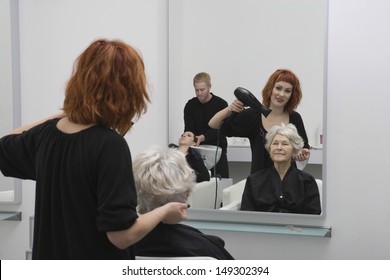 Happy Female Stylist Blow Drying Senior Woman's Hair In Salon