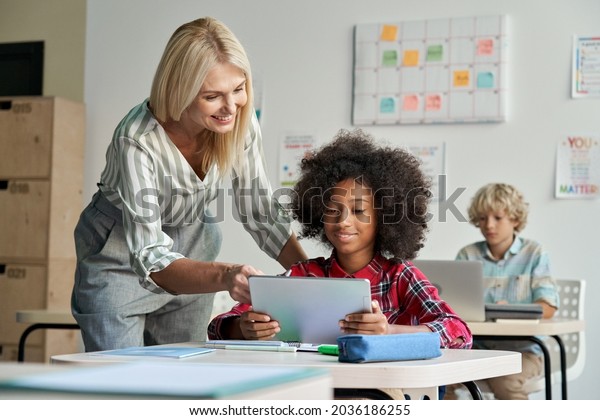 Happy female teacher tutor helping African\
American junior school kid girl student using digital tablet\
computer education program learning app technology during\
elementary class lesson in\
classroom.