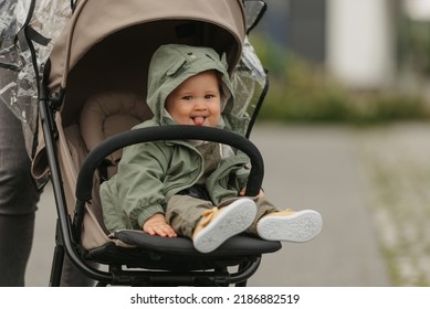 A happy female toddler with her tongue out is sitting in the stroller on a cloudy day. In a green village, a young girl in a raincoat is relaxing in a baby carriage.