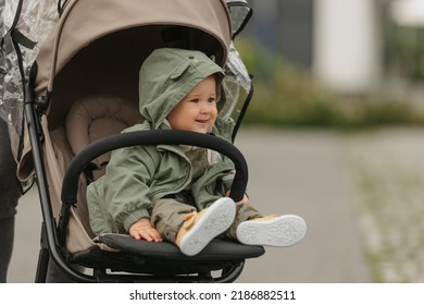 A happy female toddler is sitting in the stroller on a cloudy day. In a green village, a young girl in a raincoat is relaxing in a baby carriage.