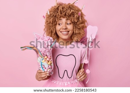 Happy female tooth fairy smiles broadly shows perfect white teeth tells what food you should avoid eating to keep them healthy has curly hair with toothbrushes stuck in it isolated on pink background