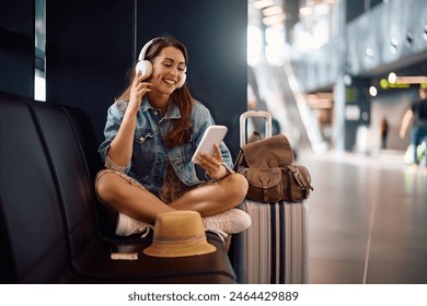 Happy female tourist listening music on headphones while using smart phone at departure area. Copy space. 