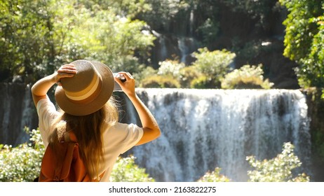 Happy female tourist in trip, journey. Young woman blogger, zoomer traveler make photo for social media of amazing waterfall, nature on trendy retro camera. Wanderlust, travel concept. Cinematic shot.