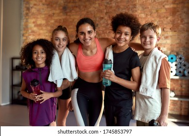 Happy female trainer and positive children smiling at camera while standing in gym. Sport, healthy lifestyle, physical education concept. Horizontal shot. Front view