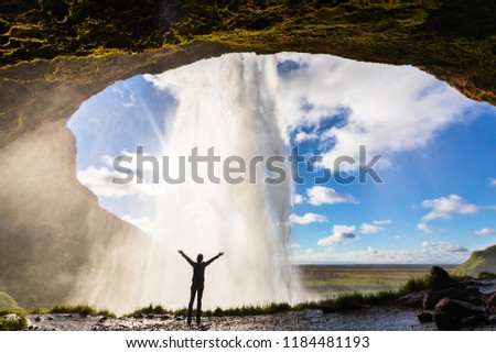 Happy female traveler feeling the power of Seljalandsfoss waterfall in the South of Iceland, person standing behind the stream, famous Icelandic landmark