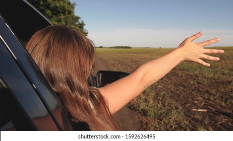 Happy Female Traveler Rides Car Along A Country Road. Hand From The Car Window Catches Wind. Girl With Long Hair Is Sitting In Front Seat Of Car, Stretching Her Arm Out Window And Catching Glare Sun