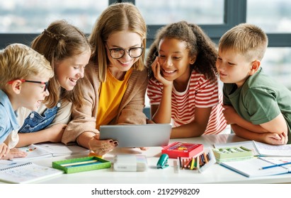 Happy female tutor and optimistic children smiling and watching video on tablet while gathering around table during lesson at school - Powered by Shutterstock