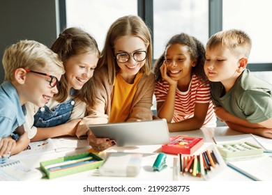 Happy female tutor and optimistic children smiling and watching video on tablet while gathering around table during lesson at school - Shutterstock ID 2199238775