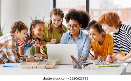 Happy female tutor and optimistic children smiling and watching laptop screen while gathering around table during lesson at school - Powered by Shutterstock