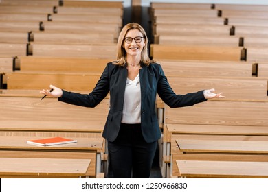 Happy Female University Professor With Outstretched Hands Looking At Camera In Classroom