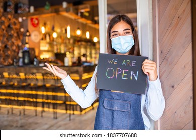 Happy Female Waitress With Protective Face Mask Holding Open Sign While Standing At Cafe Or Restaurant Doorway, Open Again After Lock Down Due To Outbreak Of Coronavirus Covid-19