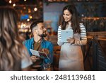 A happy female waitress wearing an apron interacts with dining guests, taking orders on a digital tablet in a cozy restaurant setting.