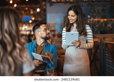 A happy female waitress wearing an apron interacts with dining guests, taking orders on a digital tablet in a cozy restaurant setting. - Powered by Shutterstock