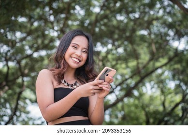 A happy Filipina woman smiling for the camera while chatting on her cellphone. Outdoor nature park scene. - Shutterstock ID 2093353156