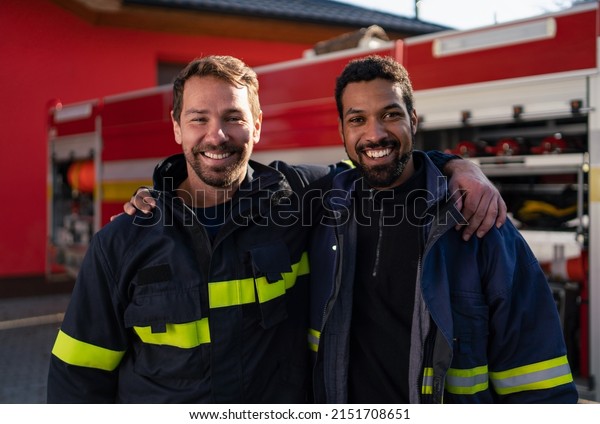 Happy firefighters crew with fire station and\
truck in background looking at\
camera.