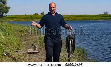 happy fisherman holds a lot of caught pike on a kukan and a metal net with fish on a background of blue lake and sky