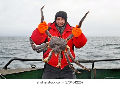 Happy fisherman in a red jacket caught and holds in his hands a beautiful huge red king crab against the backdrop of the cold North Sea, Barents sea
