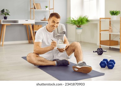 Happy fit handsome man having snack after fitness workout, sitting on mat on floor at home with dumbbells beside, holding bowl, eating healthy food, looking at camera and smiling. Sport, diet concept - Powered by Shutterstock