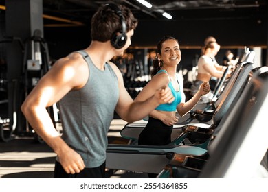 Happy fit man and woman doing cardio workout in modern gym interior, couple running on treadmills and talking with each other