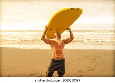 Happy Fit Senior Having Fun Surfing At Sunset Time. Sporty Bearded Man Training With Surfboard On The Beach. Elderly Healthy People Lifestyle And Extreme Sport Concept.