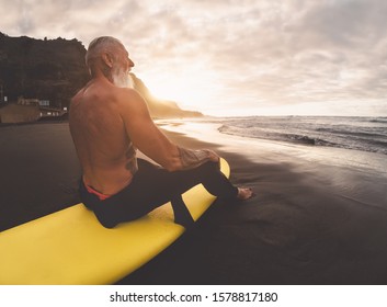 Happy fit senior man sitting on surfboard watching sunset time - Mature bearded surfer having fun on surfing day - Extreme sport and health people lifestyle concept - Powered by Shutterstock