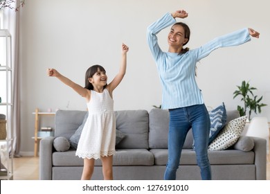 Happy Fit Sporty Mother Or Baby Sitter And Funny Active Child Daughter Having Fun Dancing Together At Home, Smiling Mom And Kid Girl Enjoy Moving To Music Raising Hands Standing In Living Room