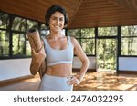 Happy fit young Hispanic woman advertising yoga class. Sporty active lady yoga coach wearing sportswear holding mat standing in yoga gym studio with big windows looking at camera. Portrait. Copy space