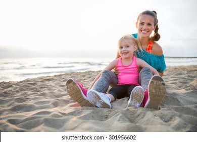 A Happy, Fit Young Mother Is Sitting With Her Young Daughter Between Her Legs. Both Are Wearing Workout Gear And Are Sitting In The Sand, Enjoying Some Rest And Relaxation After A Nice Run Together.