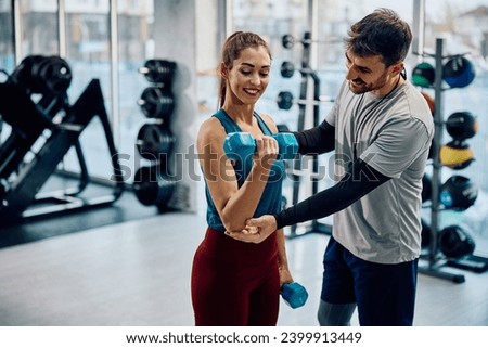 Happy fitness instructor assisting sportswoman is exercising with hand weights in a gym.