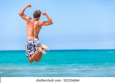 Happy Fitness Man Jumping Of Goal Achievement Success On Beach. Male Athlete Topless In Swim Shorts Doing Joy Jump On Ocean Background