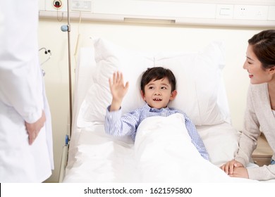 Happy Five Year Old Asian Kid Lying In Bed In Hospital Ward Waving Good-bye To Doctor