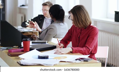 Happy Focused Business Woman In Red Dress Very Busy With Paperwork, Writing On Documents At Modern Office Table.