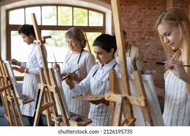 Happy focused diverse multiracial people in aprons using artist tools, enjoying painting group class drawing with paintbrushes on canvas, working together in modern studio, creative hobby activity.