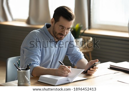 Happy focused young 30s businessman holding smartphone in hands, writing notes in personal daily planner, planning workday, checking schedule or handwriting important information at home office.