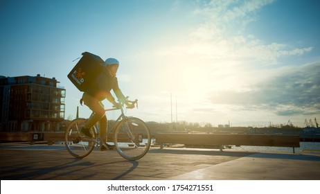 Happy Food Delivery Courier Wearing Thermal Backpack Rides a Bike on the Road To Deliver Orders and Packages for Customers. Sunny Day in City with Stylish Buildings Near Sea.