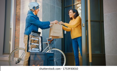 Happy Food Delivery Man Wearing Thermal Backpack On A Bike Delivers Restaurant Order To A Beautiful Female Customer. Courier Delivers Takeaway Lunch To Gorgeous Girl In Urban Building.