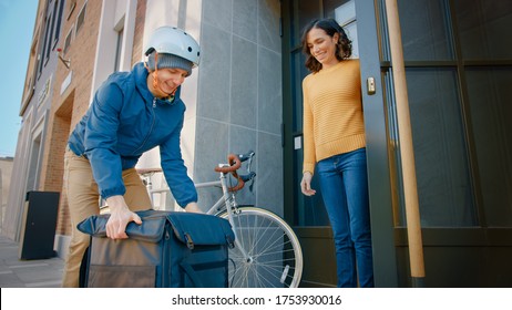 Happy Food Delivery Man Wearing Thermal Backpack On A Bike Delivers Restaurant Order To A Female Customer. Courier Delivers Takeaway Lunch To Gorgeous Girl In Modern City District Office Building