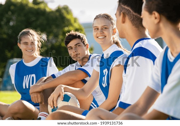 Happy football players sitting on grass on\
playground field before match. Mixed school soccer players talking\
during break. Guys and girls teammates sitting in a row for\
physical education\
lesson.
