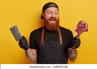 Happy Foxy Bearded Man Sells Meat With Bone In Butcher Shop, Cuts With Cleaver On Pieces, Wears Black Cap, Gloves And Apron, Isolated Over Yellow Wall. People, Production, Nutrition Concept.