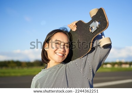 Happy and free asian girl holding cruiser board on shoulders and walking towards camera on empty road, skating on longboard and enjoying sunny weather.