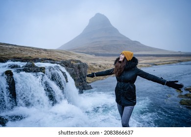 Happy free beautiful girl dancing with open arms of happiness in the wind Iceland nature. In background on famous Kirkjufell Mountain with colorful, dramatic sky.Europe travel tourist enjoying wint