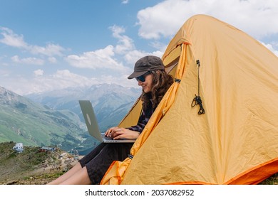 A happy and free freelance woman with glasses is typing on a laptop sitting in an orange tent in a camping high in the mountains in summer against the background of a valley and a blue sky - Powered by Shutterstock