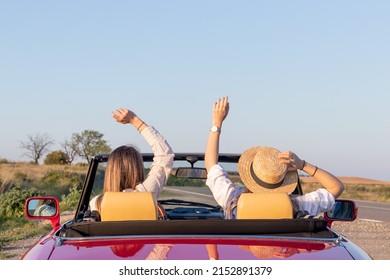 Happy Free Girlfriends With Hat Driving In Red Retro Car Cheering Joyful With Arms Raised. Road Trip Travel Concept