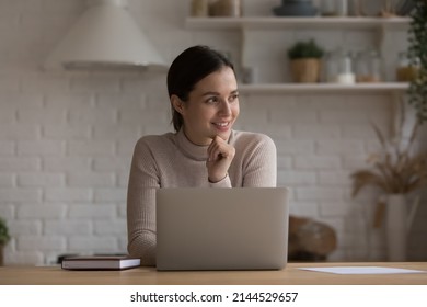 Happy freelance employee woman working at home kitchen, using laptop, thinking over online project future vision, job success, achieve, high result, smiling at good thoughts
