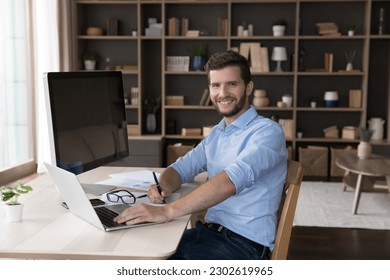 Happy freelance professional programmer sitting at table with workstation computer, typing on laptop, posing at home work table, looking at camera, smiling. Young freelancer portrait - Powered by Shutterstock