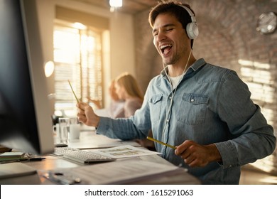 Happy freelance worker having fun while working at office desk and listening music over headphones. His colleagues are in the background. 