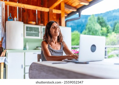 Happy freelancer woman working on laptop from the balcony of a mountain chalet, enjoying freedom and nature during summer vacation - Powered by Shutterstock