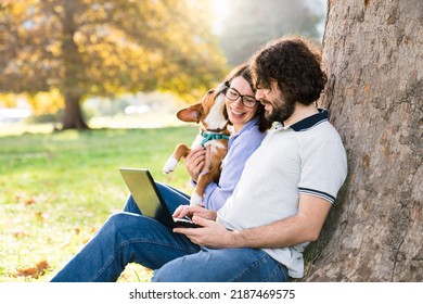 Happy Freelancers And Dog Sit With Laptop In Autumn Park Under Tree. People And Puppy Having Fun Together Outdoors. Pet Owners Enjoying Working, Shopping Online In Nature. Authentic Lifestyle